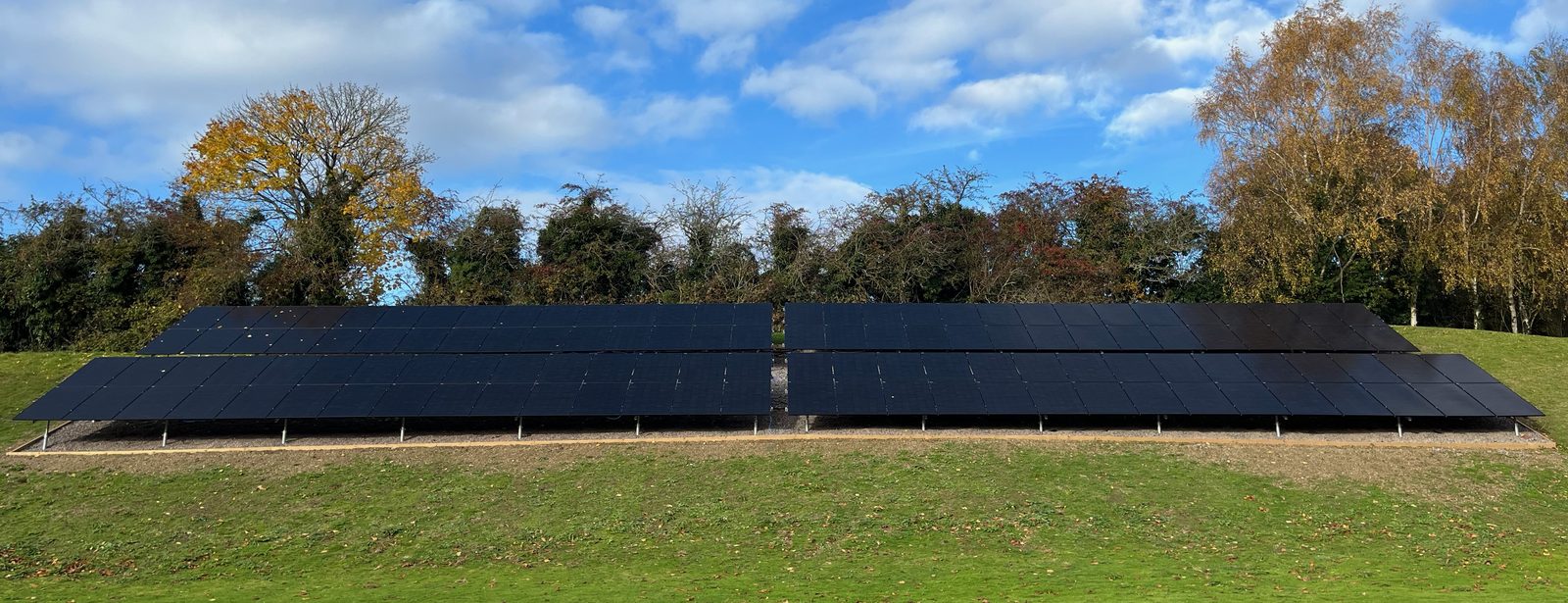 A completed ground mounted solar array installed by Sy the Solar Guy in Surrey, two long rows of black panels in a field backed by autumn trees